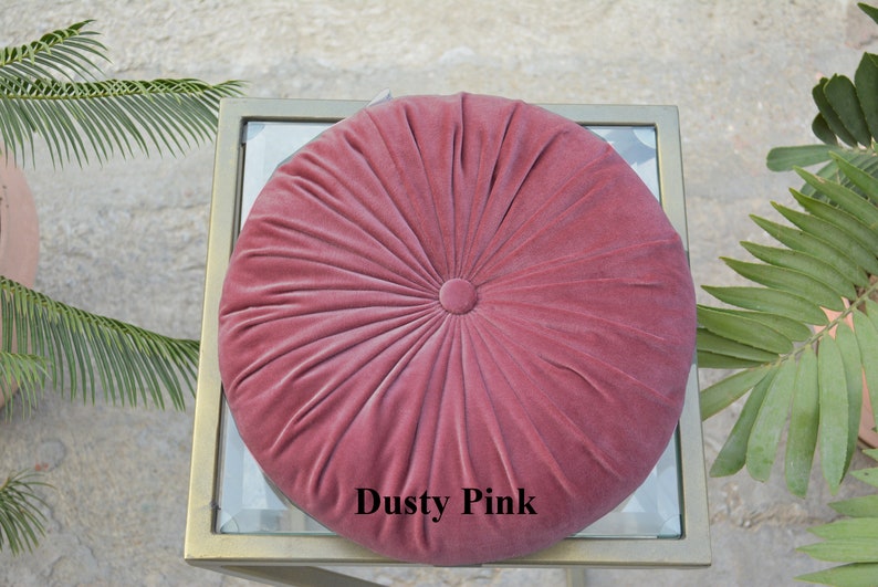 May include: A round, dusty pink velvet cushion with a button center. The cushion has a radial design and is placed on a glass-topped table with a gold frame. The text "Dusty Pink" is visible on the cushion.