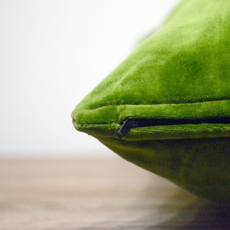 May include: A close-up of a lime green velvet cushion. The cushion has a black zip closure and is on a light brown surface. The cushion is a square shape and has a soft, plush texture.