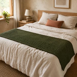 Puede incluir: Un camino de cama rectangular de color verde oliva oscuro con textura, hecho de un material afelpado y suave, colocado horizontalmente sobre una cama con ropa de cama de lino beige claro, dos almohadas a juego y un cojín rectangular de color terracota.