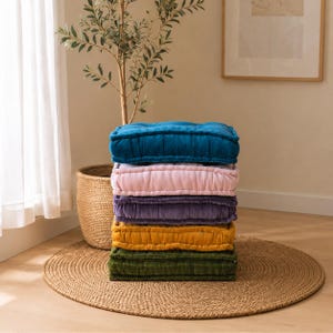Puede incluir: Una pila de cinco cojines de suelo cuadrados de tela de terciopelo, con una vibrante paleta de colores en azul cerceta, rosa claro, morado, amarillo mostaza y verde oliva oscuro. Cada cojín tiene una superficie capitoné con bordes cosidos.