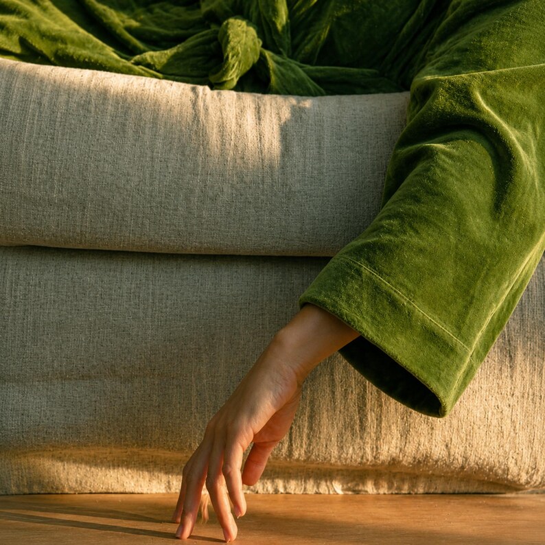 May include: A plush, olive green velvet robe with long sleeves and a tie-waist closure, draped over a light beige, textured linen-upholstered armchair. The fabric of the robe shows a soft, light-reflecting pile, while the chair features a woven texture.