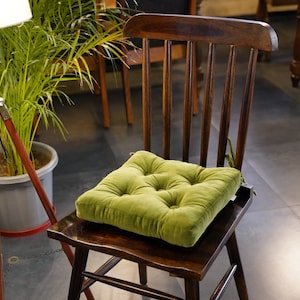 Puede incluir: Una silla de madera oscura con un cojín verde. El cojín cuadrado es acolchado y tiene una textura aterciopelada. La silla tiene un respaldo curvo con barrotes verticales. Una lámpara de pie y una planta en maceta están en el fondo.
