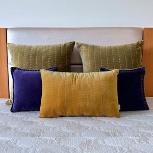 Op de afbeelding: Een verzameling rechthoekige kussens in verschillende kleuren. De kussens zijn in olijfgroen, mosterdgeel en diep paars. Ze liggen op een bed met een wit dekbed met patroon.