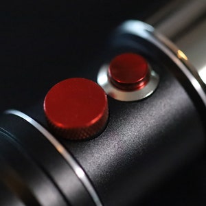 May include: Close-up of a black lightsaber hilt with two red buttons. The buttons are round and have a silver ring around them.