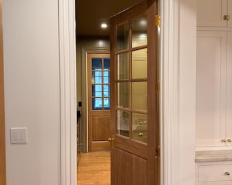 May include: A partially open wooden door with glass panes, revealing a glimpse into another room. The door has a warm, natural wood finish and brass hardware. The interior features hardwood floors and a second wooden door in the distance.