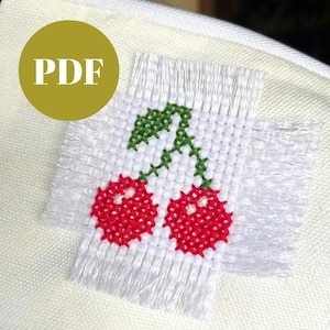 May include: A white cross-stitch design featuring two red cherries with green leaves and stems. The design is on a white fabric with fringed edges. A gold circle with the text "PDF" is in the upper left corner.