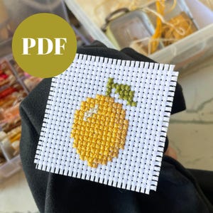 May include: A square cross-stitch pattern featuring a yellow lemon with a green stem and leaves. The design is on a white background with black grid lines. A yellow circle with the word "PDF" is in the upper left corner.