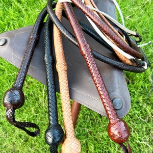 May include: A collection of braided leather whips in various colors, including brown, black, and tan. The whips are arranged on a brown leather surface and have a braided handle with a rounded end.