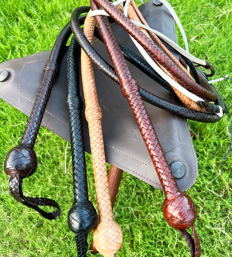 May include: A collection of braided leather whips in various colors, including brown, black, and tan. The whips are arranged on a brown leather surface and have a braided handle with a rounded end.