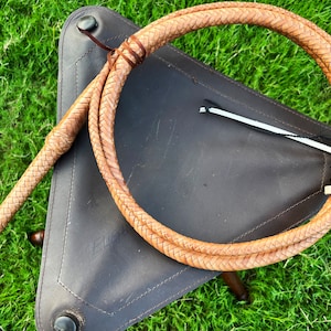 May include: A braided leather bullwhip with a tan color and a knotted handle. The whip is coiled on a brown leather surface.