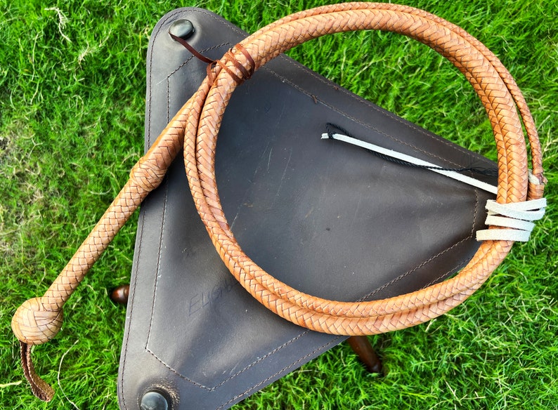 May include: A braided leather bullwhip with a tan color and a knotted handle. The whip is coiled on a brown leather surface.