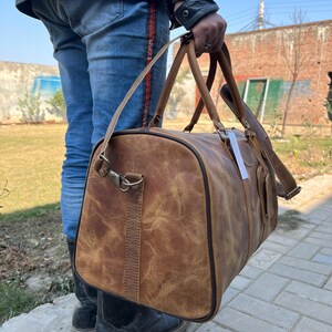 K&ouml;nnte beinhalten: Eine braune Reisetasche aus Leder mit Used-Look, ausgestattet mit zwei Tragegriffen, einem abnehmbaren Schultergurt mit Metallbeschl&auml;gen, einem seitlichen Rei&szlig;verschluss und einem kleinen wei&szlig;en rechteckigen Etikett am Griff.