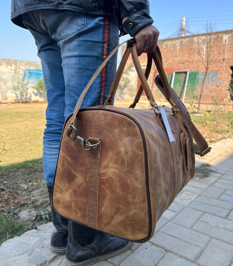 K&ouml;nnte beinhalten: Eine braune Reisetasche aus Leder mit Used-Look, ausgestattet mit zwei Tragegriffen, einem abnehmbaren Schultergurt mit Metallbeschl&auml;gen, einem seitlichen Rei&szlig;verschluss und einem kleinen wei&szlig;en rechteckigen Etikett am Griff.