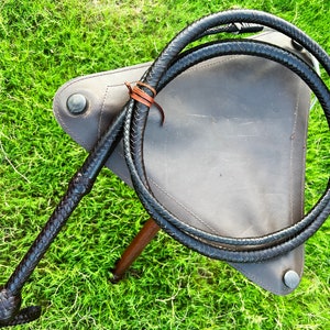 May include: A black leather bullwhip with a braided handle. The whip is coiled and resting on a brown leather surface.