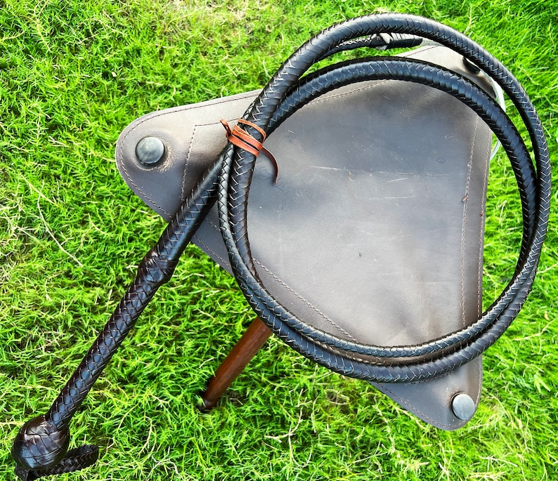 May include: A black leather bullwhip with a braided handle. The whip is coiled and resting on a brown leather surface.