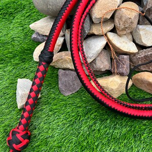 May include: A red and black braided bullwhip with a knotted handle. The whip is lying on a bed of green grass and rocks.