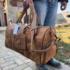 K&ouml;nnte beinhalten: Eine hellbraune Reisetasche aus Leder mit Used-Look, ausgestattet mit zwei Tragegriffen, einem abnehmbaren Schultergurt, einem seitlichen Rei&szlig;verschlussfach und einem rechteckigen Kofferanh&auml;nger mit einem wei&szlig;en Papieretikett.
