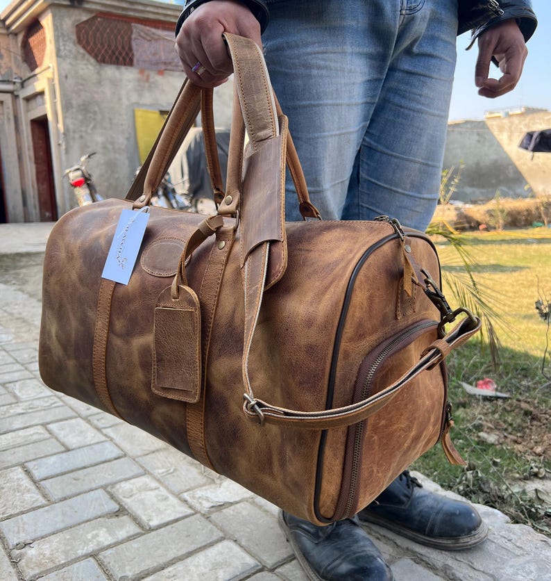 K&ouml;nnte beinhalten: Eine hellbraune Reisetasche aus Leder mit Used-Look, ausgestattet mit zwei Tragegriffen, einem abnehmbaren Schultergurt, einem seitlichen Rei&szlig;verschlussfach und einem rechteckigen Kofferanh&auml;nger mit einem wei&szlig;en Papieretikett.