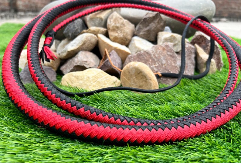 May include: A red and black braided leather bullwhip. The whip is coiled and lying on a bed of green grass.