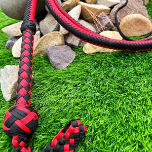 May include: A black and red braided leather whip with a knotted handle. The whip is lying on a bed of green artificial grass.