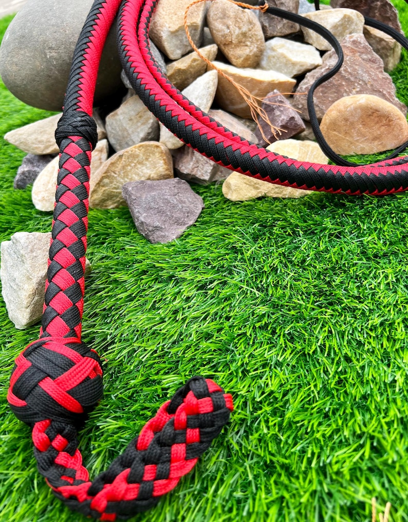 May include: A black and red braided leather whip with a knotted handle. The whip is lying on a bed of green artificial grass.