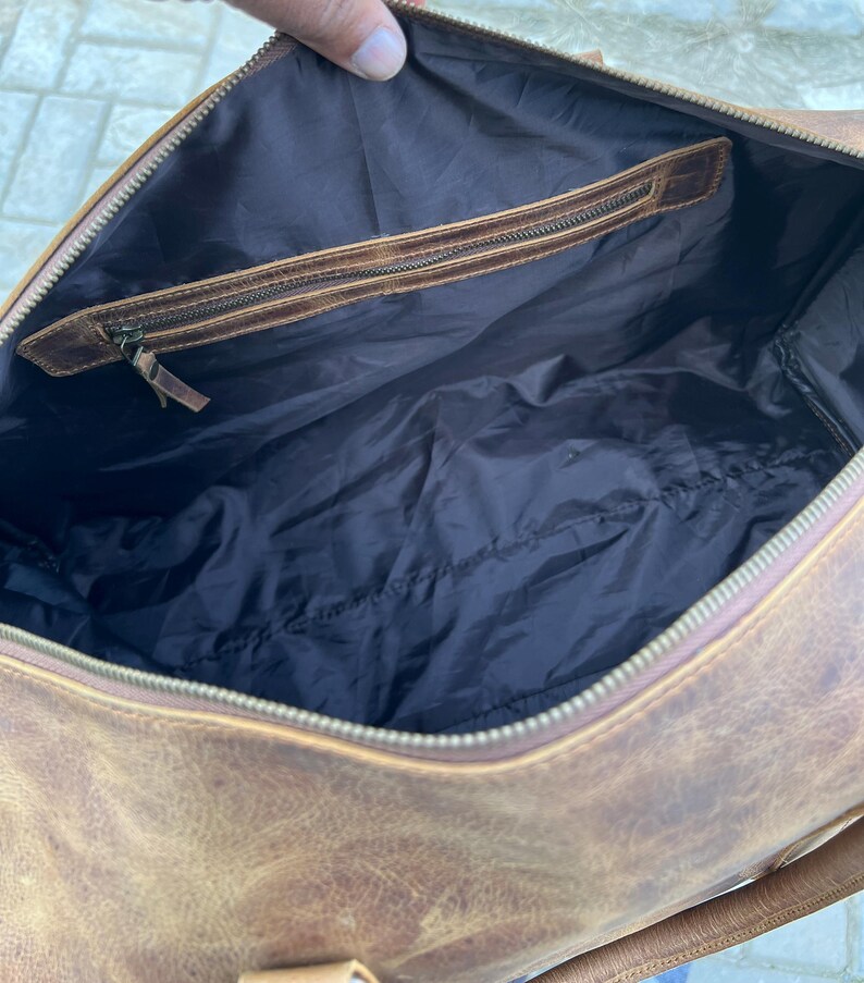 K&ouml;nnte beinhalten: Eine braune Reisetasche aus Leder mit dunkelbraunem Innenfutter. Die Tasche verf&uuml;gt &uuml;ber einen Rei&szlig;verschluss an der Oberseite und eine Innentasche mit Rei&szlig;verschluss und Lederbesatz. Das Au&szlig;enleder hat eine strukturierte, abgenutzte Optik.