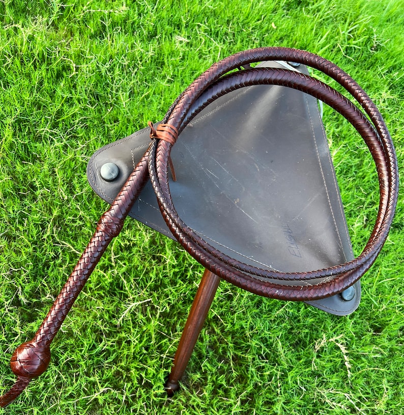 May include: A brown leather bullwhip with a braided handle. The whip is coiled around a brown leather stool.