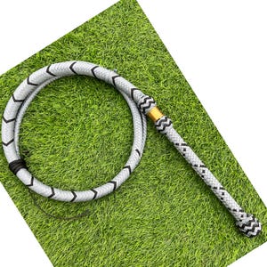 May include: A braided leather whip in a light gray color with black chevron and diamond patterns. The handle features a gold-colored metal accent and a black and white woven design. The whip is coiled on a green surface.