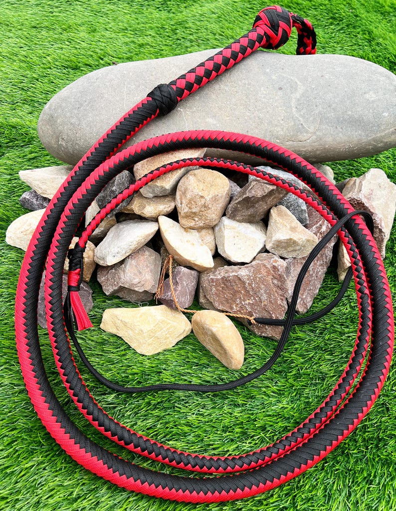 May include: A black and red braided bullwhip with a knotted handle. The whip is coiled on a bed of rocks and green grass.