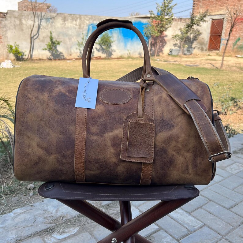 K&ouml;nnte beinhalten: Eine braune Reisetasche aus Leder mit Used-Look, ausgestattet mit zwei Tragegriffen, einem abnehmbaren Schultergurt mit Polsterung und einem passenden Leder-Gep&auml;ckanh&auml;nger. Ein kleines wei&szlig;es rechteckiges Etikett ist am Griff befestigt.
