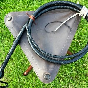 May include: A black leather bullwhip with a braided handle, coiled and resting on a brown leather stool. The whip is approximately 4 feet long.