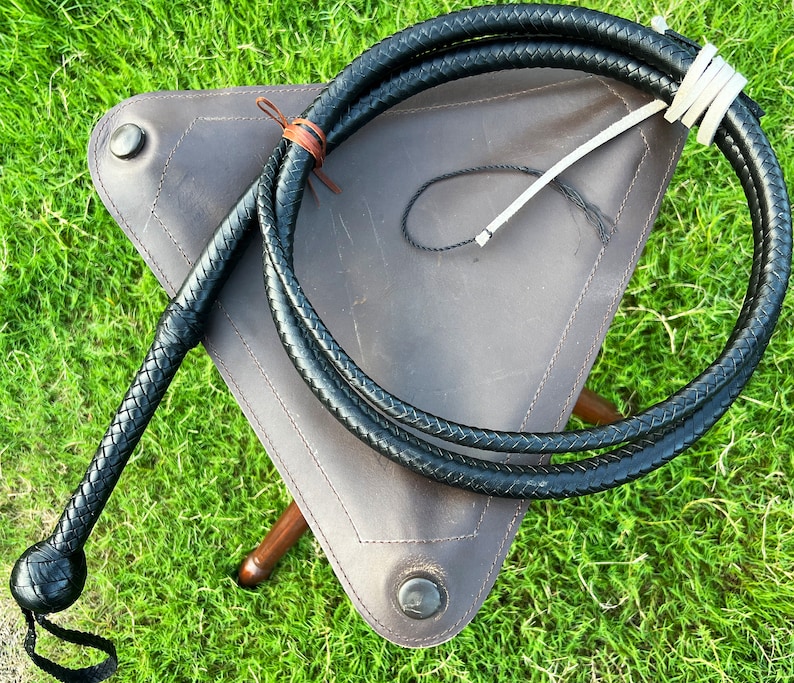 May include: A black leather bullwhip with a braided handle, coiled and resting on a brown leather stool. The whip is approximately 4 feet long.