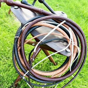 May include: A set of three braided leather whips in brown, tan, and black colors. The whips are coiled together and resting on a brown leather stool.