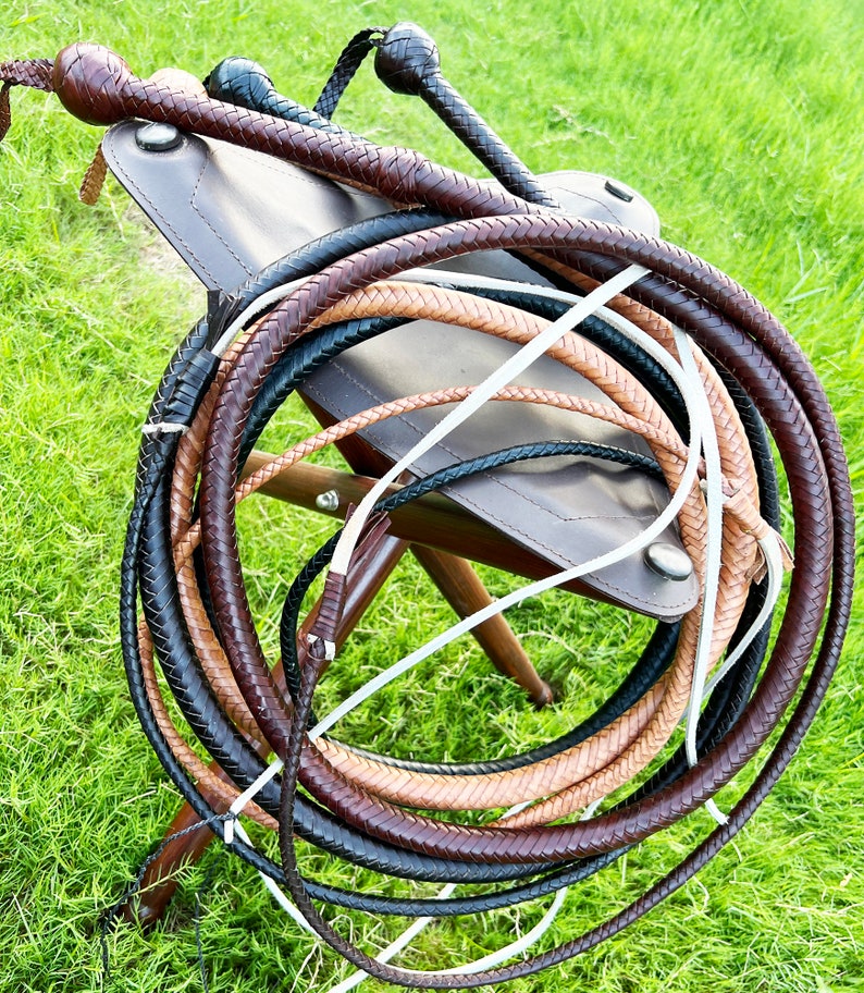 May include: A set of three braided leather whips in brown, tan, and black colors. The whips are coiled together and resting on a brown leather stool.