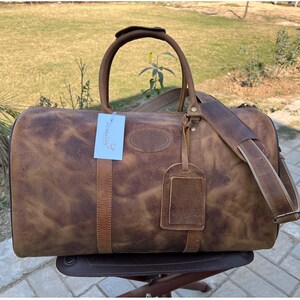 K&ouml;nnte beinhalten: Eine braune Reisetasche aus Leder mit Used-Look, ausgestattet mit zwei Tragegriffen, einem abnehmbaren Schultergurt und einem passenden Kofferanh&auml;nger. Ein wei&szlig;es rechteckiges Etikett mit blauem Text ist am vorderen Griff befestigt.