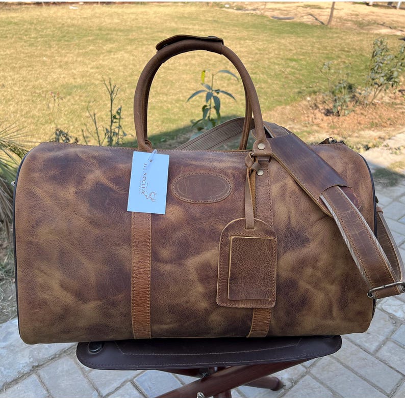 K&ouml;nnte beinhalten: Eine braune Reisetasche aus Leder mit Used-Look, ausgestattet mit zwei Tragegriffen, einem abnehmbaren Schultergurt und einem passenden Kofferanh&auml;nger. Ein wei&szlig;es rechteckiges Etikett mit blauem Text ist am vorderen Griff befestigt.