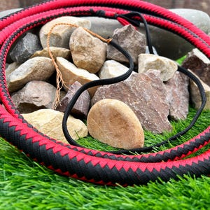 May include: A red and black braided whip with a woven design. The whip is lying on a bed of rocks and green grass.
