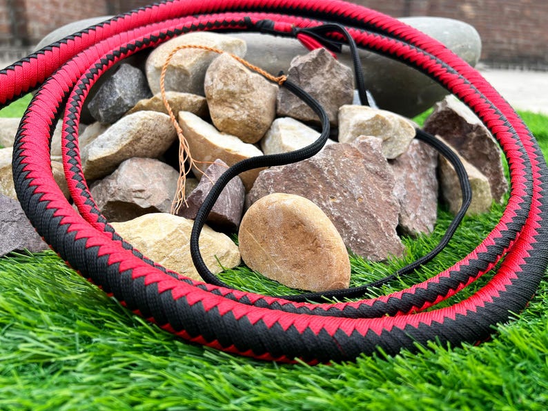 May include: A red and black braided whip with a woven design. The whip is lying on a bed of rocks and green grass.