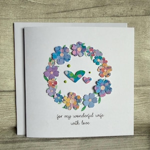 May include: A white greeting card with a floral wreath design. The wreath is composed of colorful paper flowers in shades of blue, purple, and yellow. Two hearts and the text "for my wonderful wife with love" are in the center.