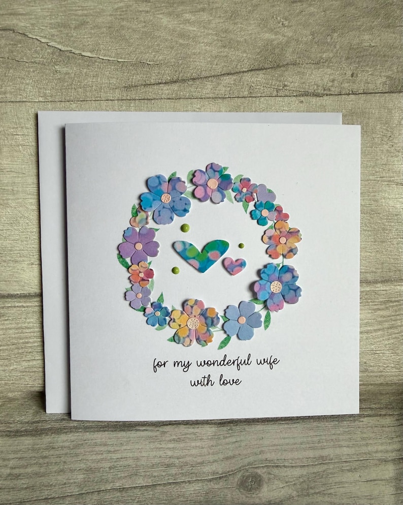May include: A white greeting card with a floral wreath design. The wreath is composed of colorful paper flowers in shades of blue, purple, and yellow. Two hearts and the text "for my wonderful wife with love" are in the center.