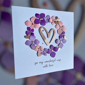 May include: A light blue greeting card with a floral wreath design. The wreath is composed of purple, pink, and gold flowers surrounding a gold heart. The text "for my wonderful wife with love" is written in black.