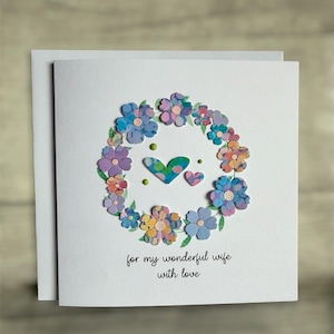 May include: A handmade greeting card with a floral wreath design. The wreath is composed of colorful paper flowers in shades of blue, purple, and yellow. Two hearts and the text "for my wonderful wife with love" are featured on the card.