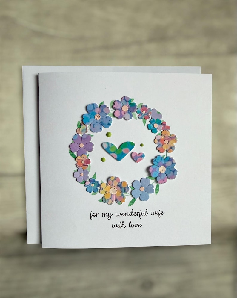 May include: A handmade greeting card with a floral wreath design. The wreath is composed of colorful paper flowers in shades of blue, purple, and yellow. Two hearts and the text "for my wonderful wife with love" are featured on the card.