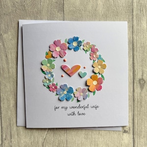 May include: A handmade greeting card with a floral wreath design. The wreath is composed of colorful paper flowers in shades of pink, blue, yellow, and orange. Two hearts are in the center. The text reads "for my wonderful wife with love."