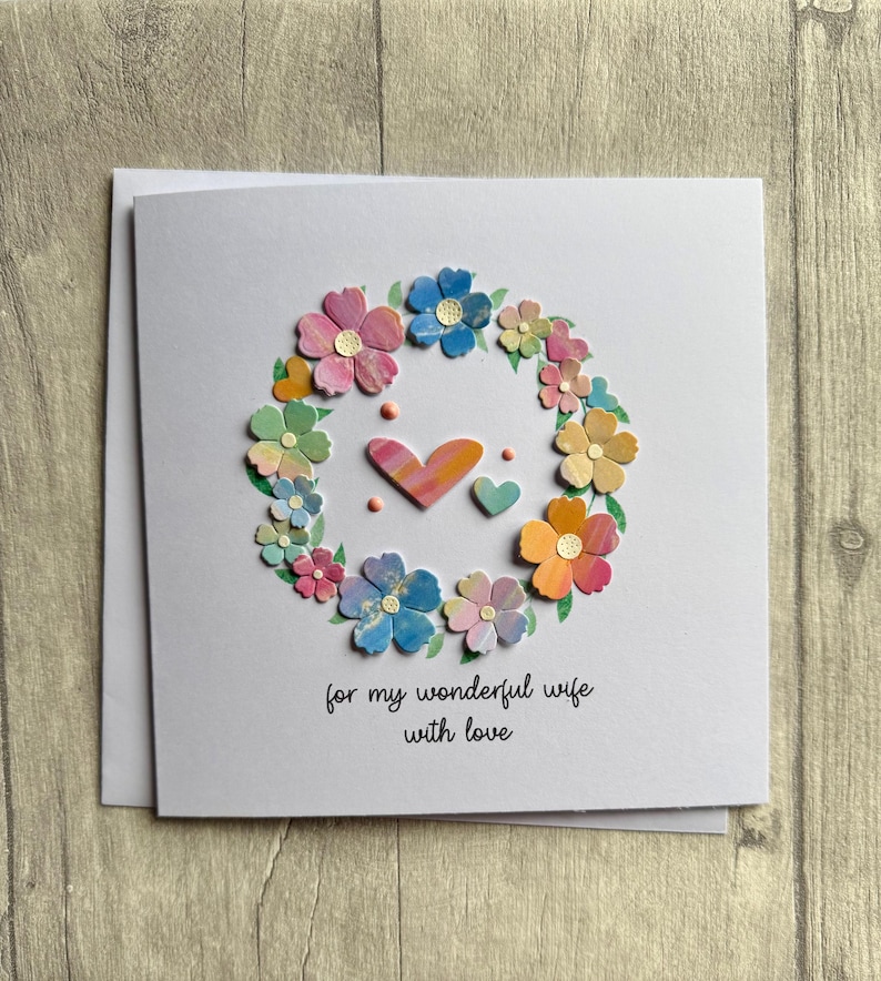 May include: A handmade greeting card with a floral wreath design. The wreath is composed of colorful paper flowers in shades of pink, blue, yellow, and orange. Two hearts are in the center. The text reads "for my wonderful wife with love."
