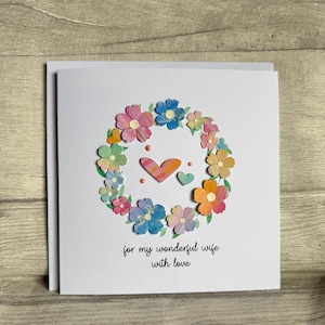 May include: A white greeting card with a floral wreath design. The wreath is composed of colorful paper flowers in shades of pink, blue, orange, and yellow. Two hearts and the text "for my wonderful wife with love" are in the center.