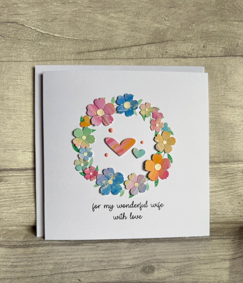 May include: A white greeting card with a floral wreath design. The wreath is composed of colorful paper flowers in shades of pink, blue, orange, and yellow. Two hearts and the text "for my wonderful wife with love" are in the center.