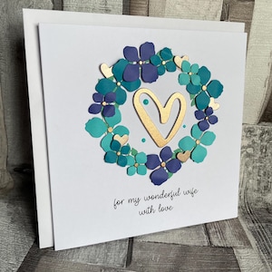 May include: A white greeting card with a floral wreath design. The wreath features teal and purple flowers with gold accents, surrounding a gold heart. The card includes the text "for my wonderful wife with love."