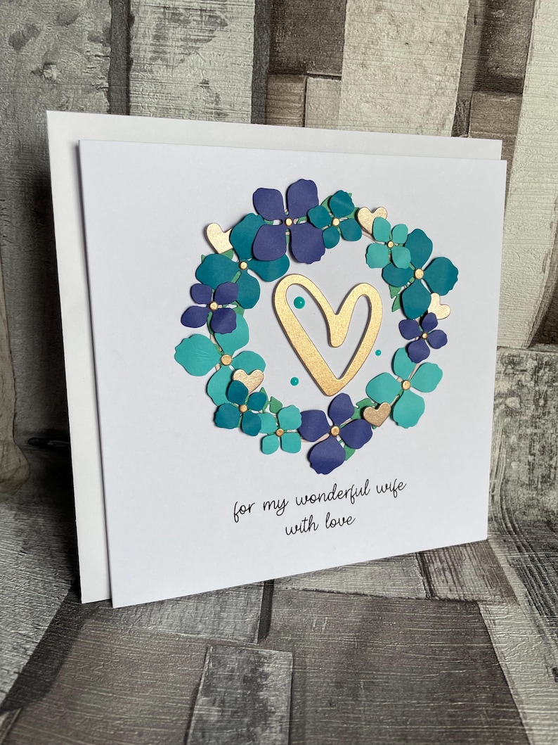 May include: A white greeting card with a floral wreath design. The wreath features teal and purple flowers with gold accents, surrounding a gold heart. The card includes the text "for my wonderful wife with love."