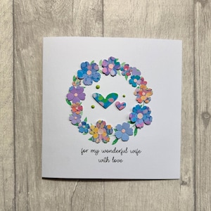 May include: A white greeting card with a floral wreath design. The wreath is composed of colorful, textured flowers in shades of blue, purple, pink, and yellow. Two hearts are in the center. The text reads, "for my wonderful wife with love."